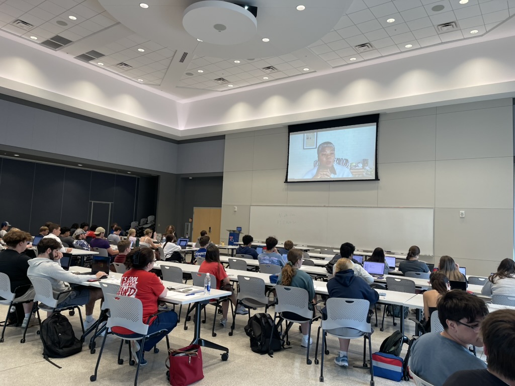 Students in a full classroom listening to a virtual panelist presented on a large screen at the front of the room.